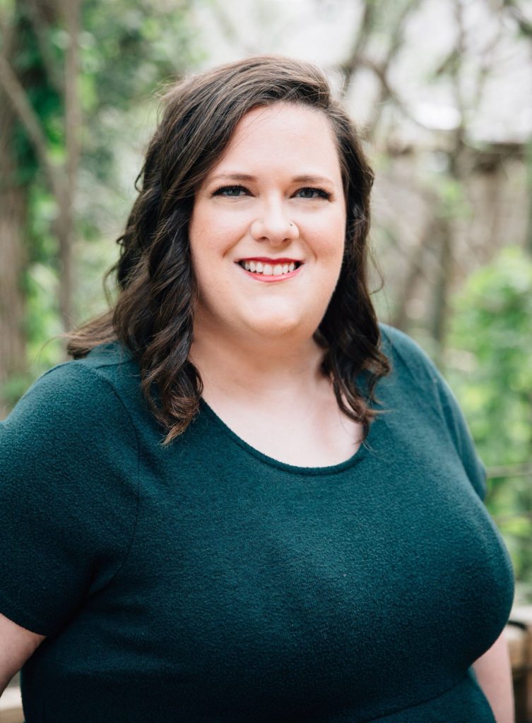 A professional photo of a white non-binary person, OB Horak, wearing a forest green dress standing in front of greenery.