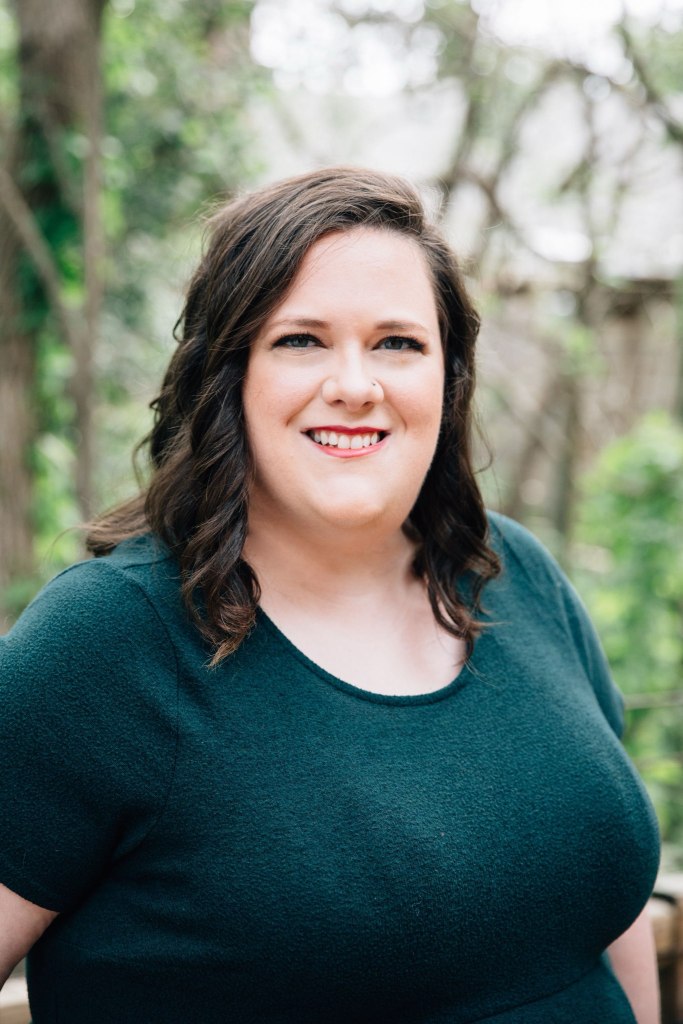 A professional photo of a white non-binary person, OB Horak, wearing a forest green dress standing in front of greenery.