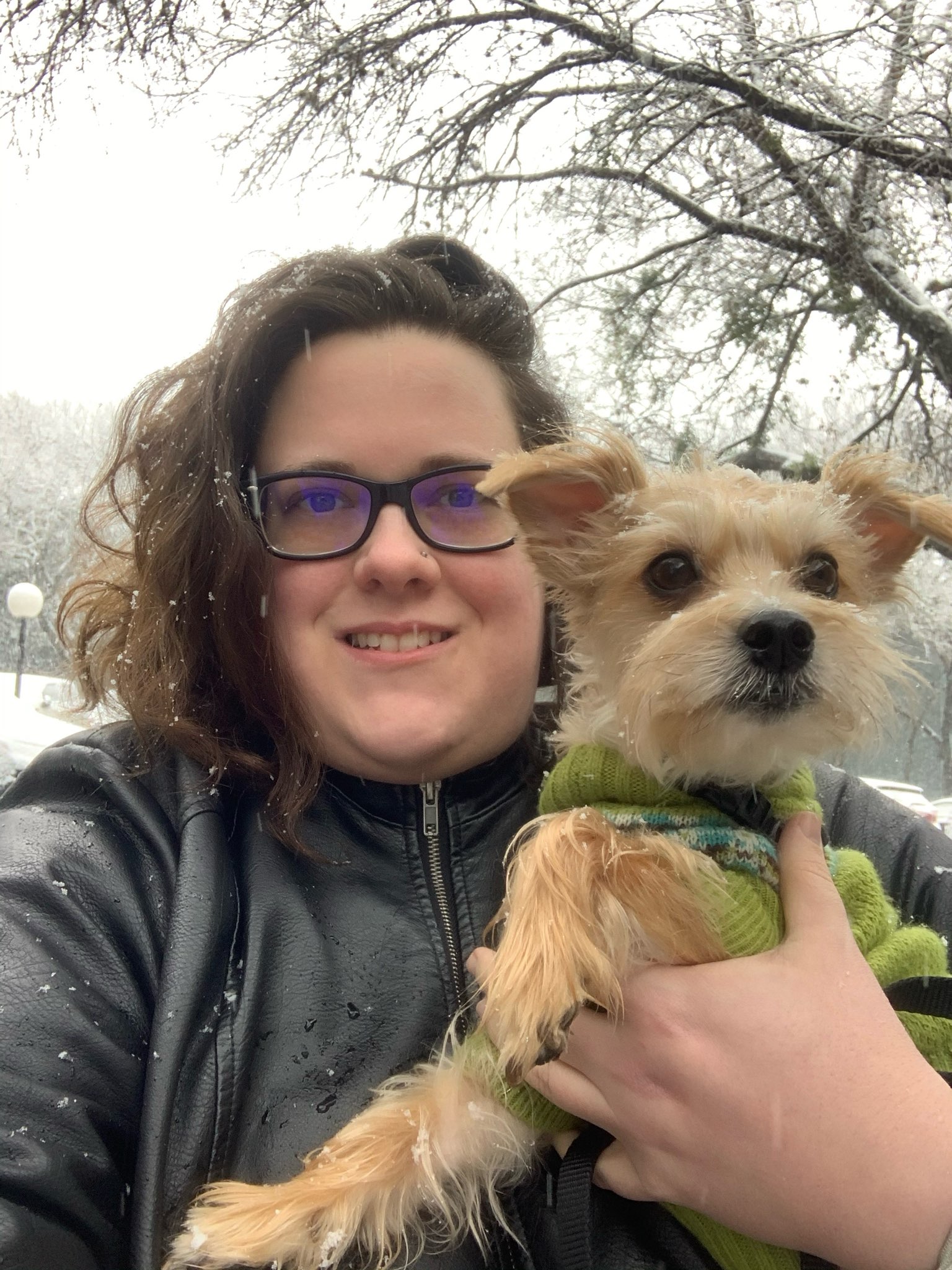 A picture of a white non-binary person, OB Horak, holding Cody Little, a mixed Yorkie Maltese wearing a green sweater on a snowy day in Austin TX.