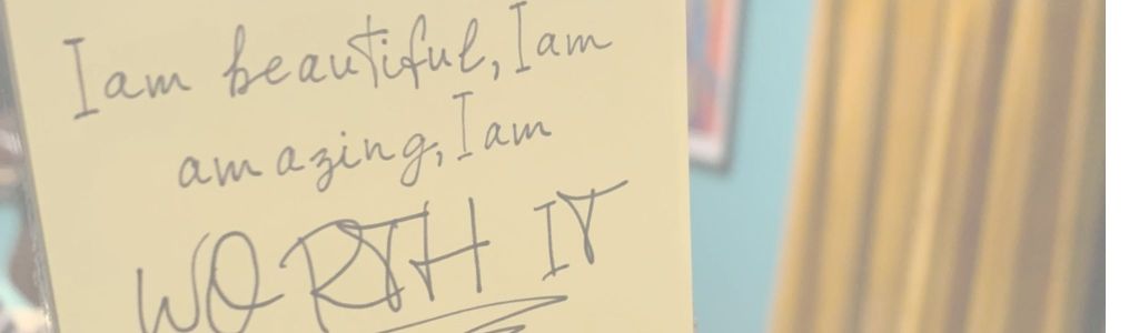 Yellow Post-it note on a mirror with the words "I am beautiful, I am amazing, I am WORTH IT," displayed at Meow Wolf Grapevine's The Real Unreal exhibition.