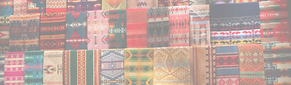 Approximately 40 Mexican art blankets displayed in three rows, each showcasing bold, bright colors and intricate woven patterns with rich textures.