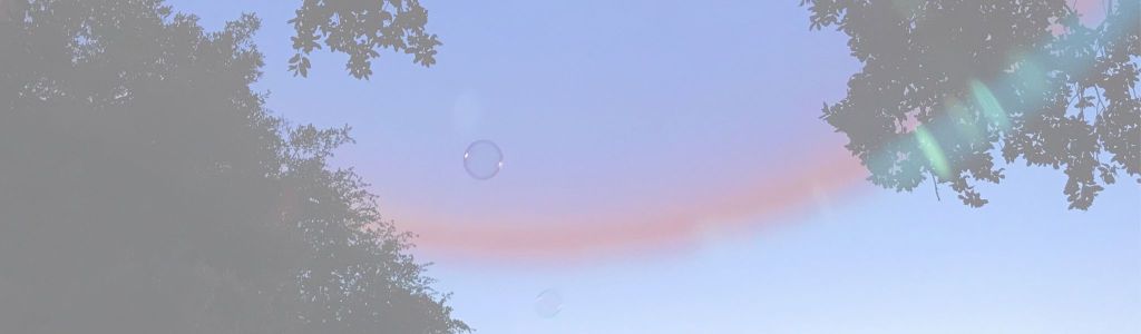 Two small, iridescent bubbles trapped inside a larger bubble close to the camera, reflecting light and hints of the surroundings, with green trees and a bright blue sky in the background. Photo taken at Austin Queer Bomb 2018.